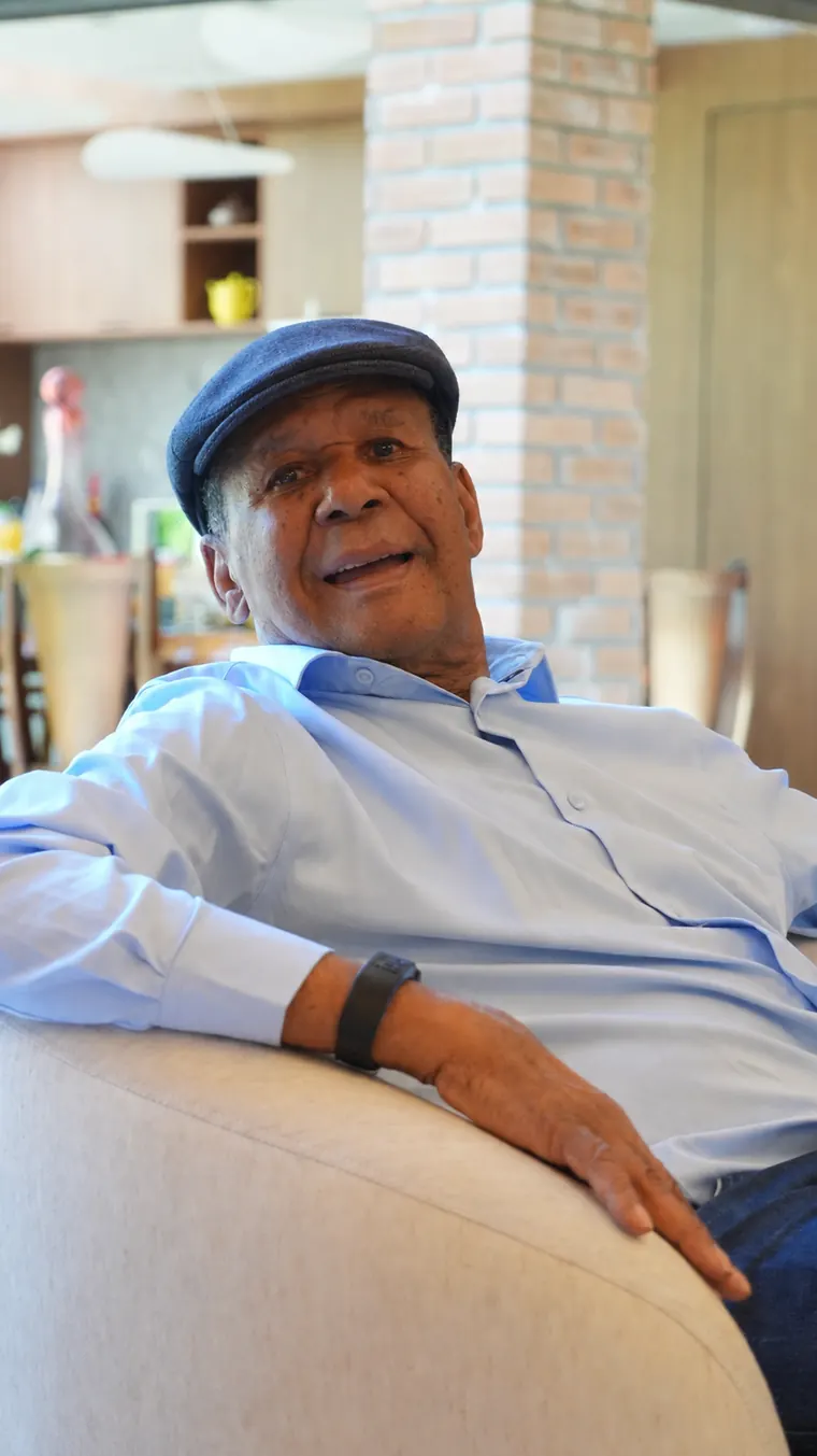 João Evangelista, de boina, acomodado em um sofá, sorrindo